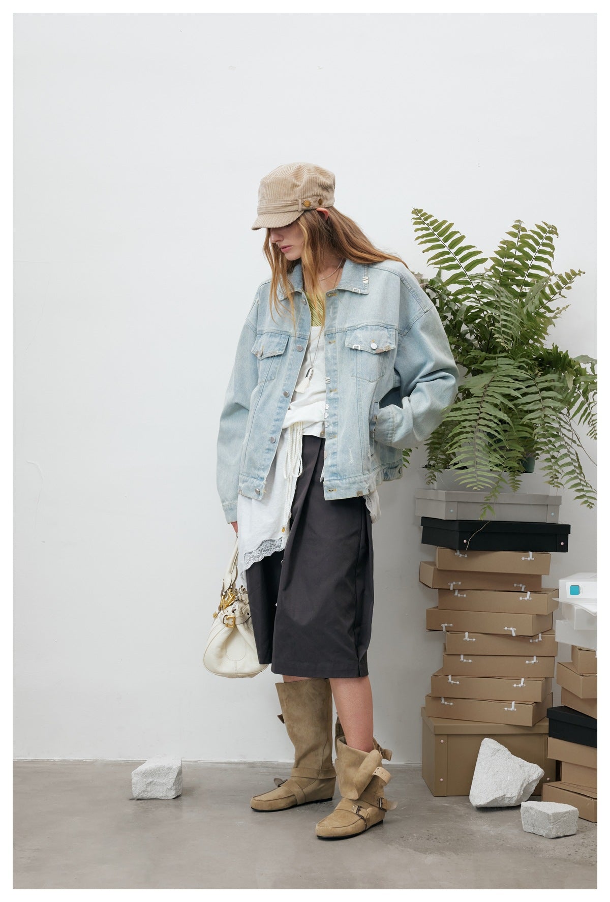 Model wearing a light blue denim jacket in a three-quarter pose with her head tilted down, styled with a cap and boots