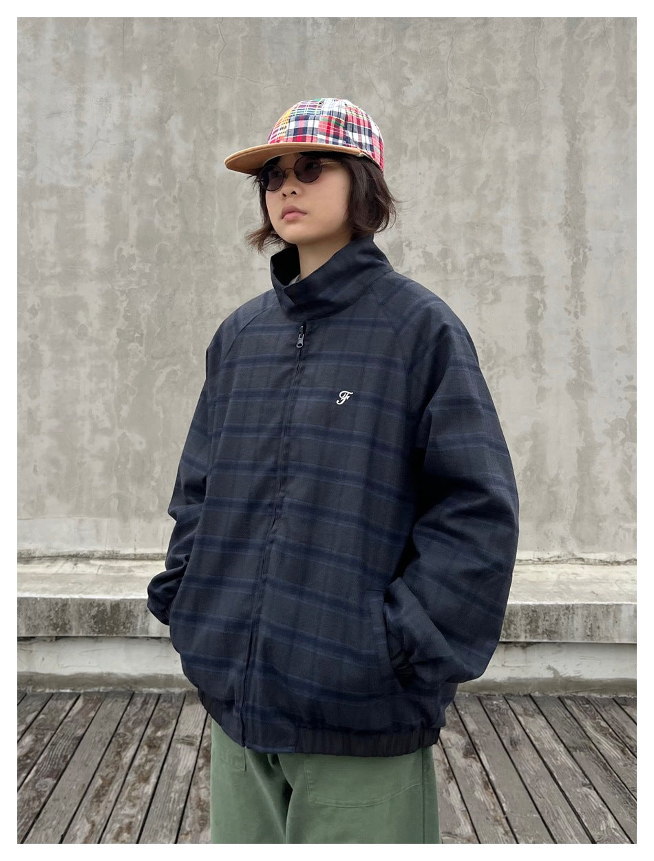 Person wearing a navy plaid stand-collar zip jacket with a small script F logo, hands in pockets, patchwork cap