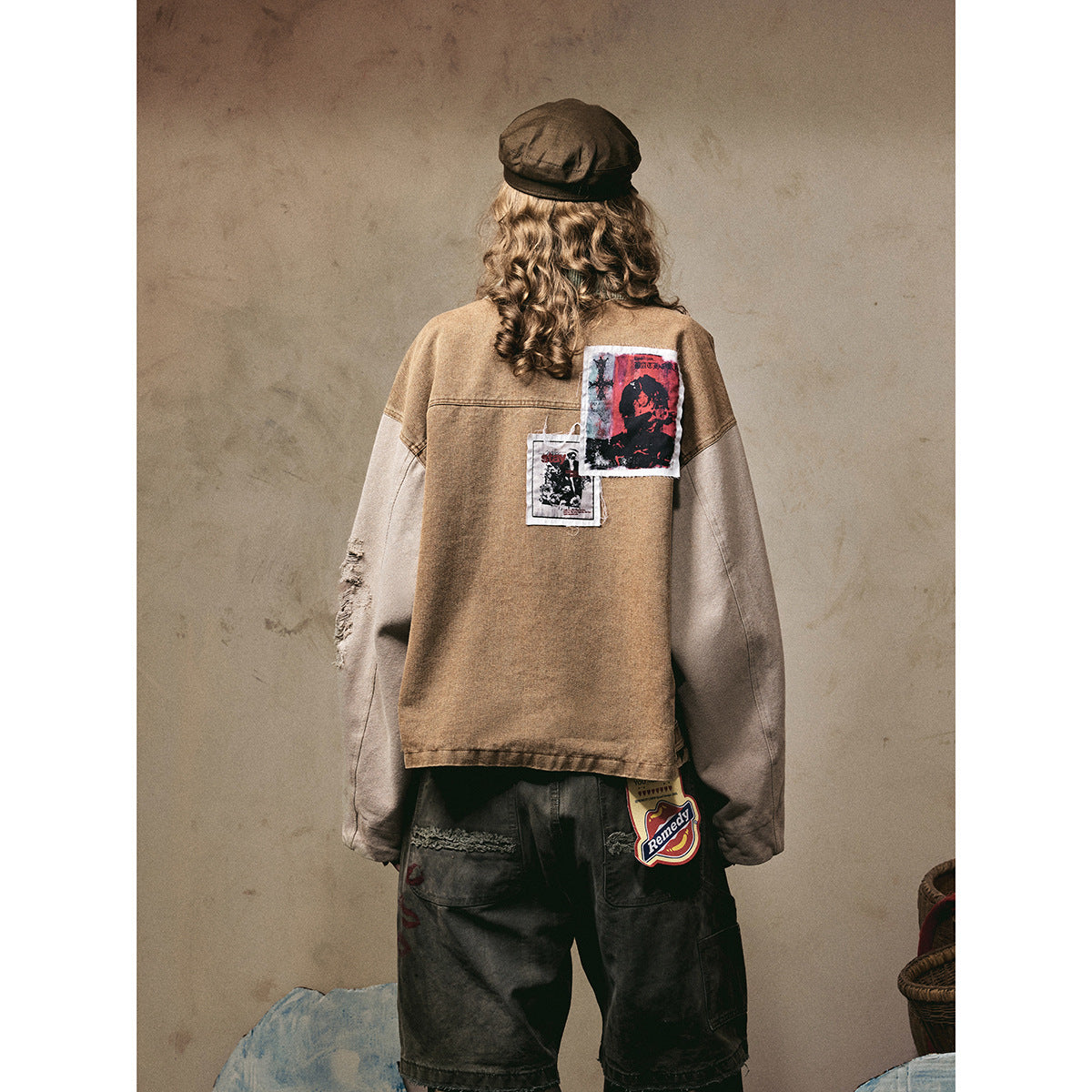 Back view of a model wearing a brown jacket with pale sleeves and graphic patches, with loose dark pants.