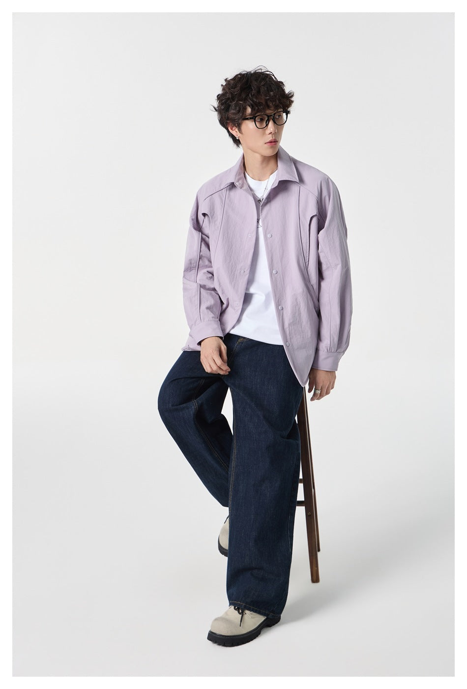 Model seated on a stool wearing an open pale lavender shirt over a white tee and dark wide-leg jeans.
