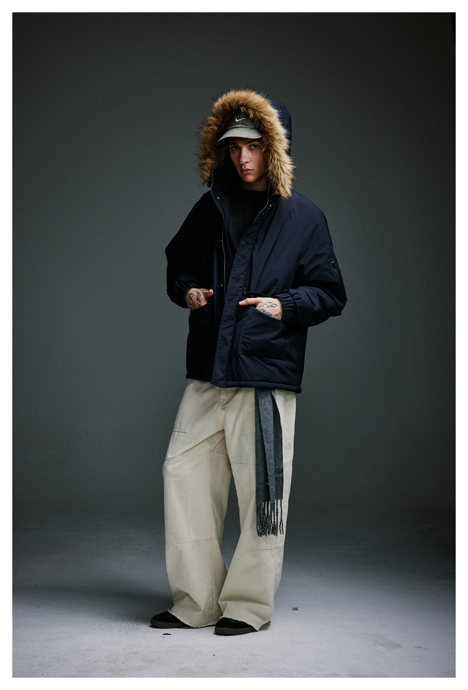 Model wearing a navy fur-trimmed parka with hands in its front pockets and beige wide-leg trousers.