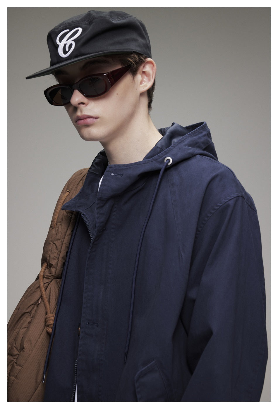 Close-up of a model wearing a navy hooded jacket with drawstring hood, cap and sunglasses