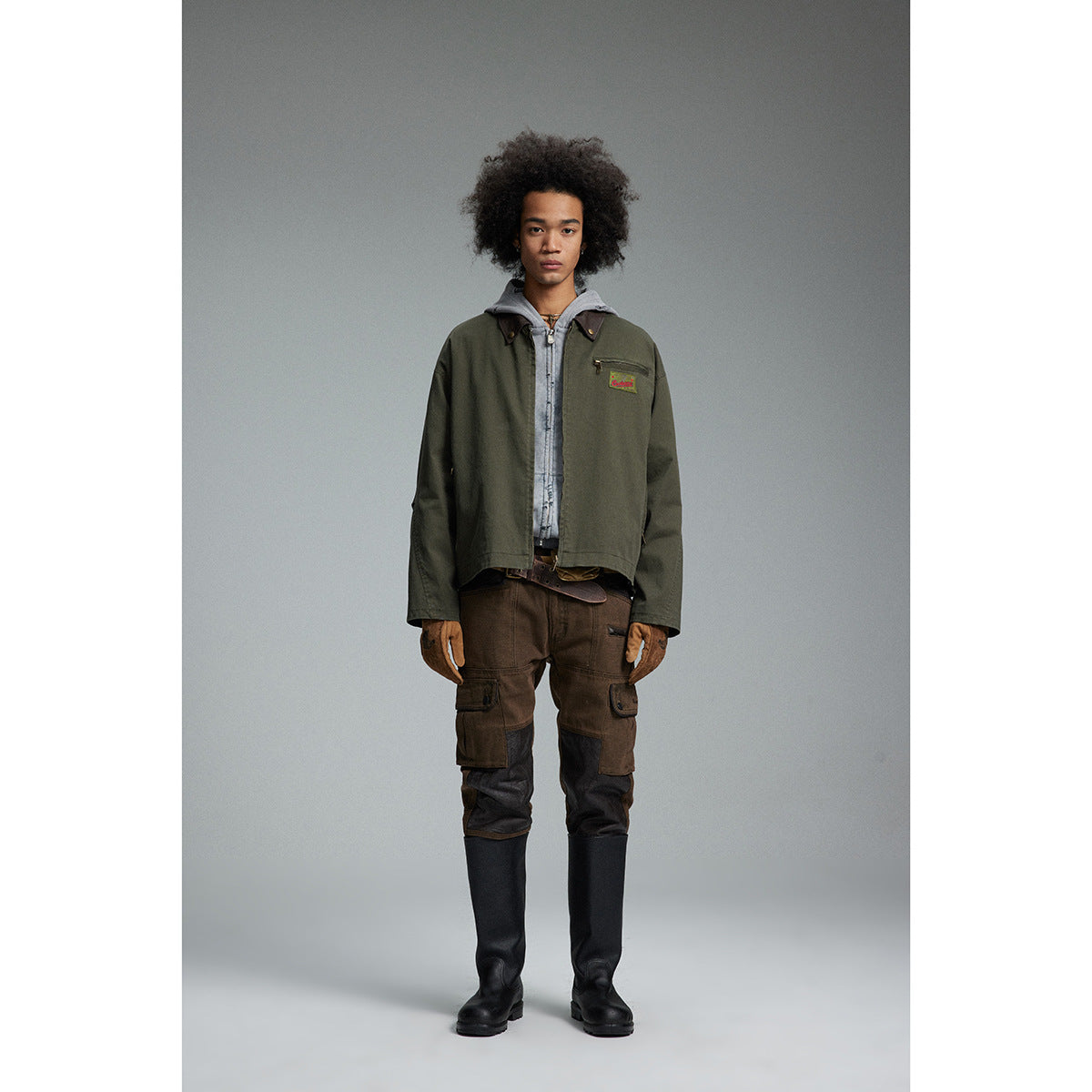 Full-length model wearing an olive work jacket over a gray hoodie, brown cargo pants, gloves and black boots.