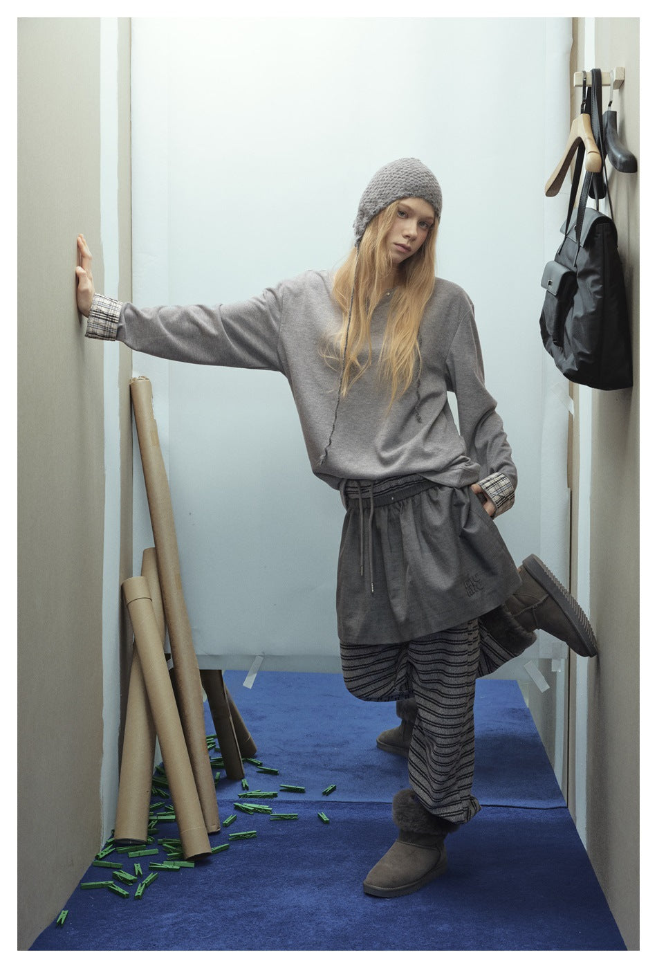 Model wearing a gray pullover with plaid cuffs, dark drawstring shorts over striped trousers, and a gray knit hat.