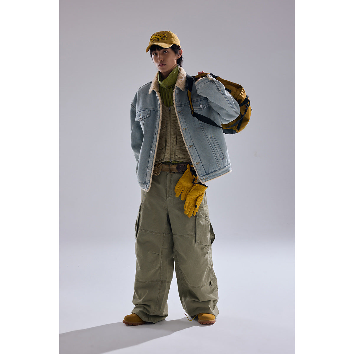 Model wears a light blue sherpa-lined denim jacket with olive cargo pants, a yellow cap and gloves, and a backpack.
