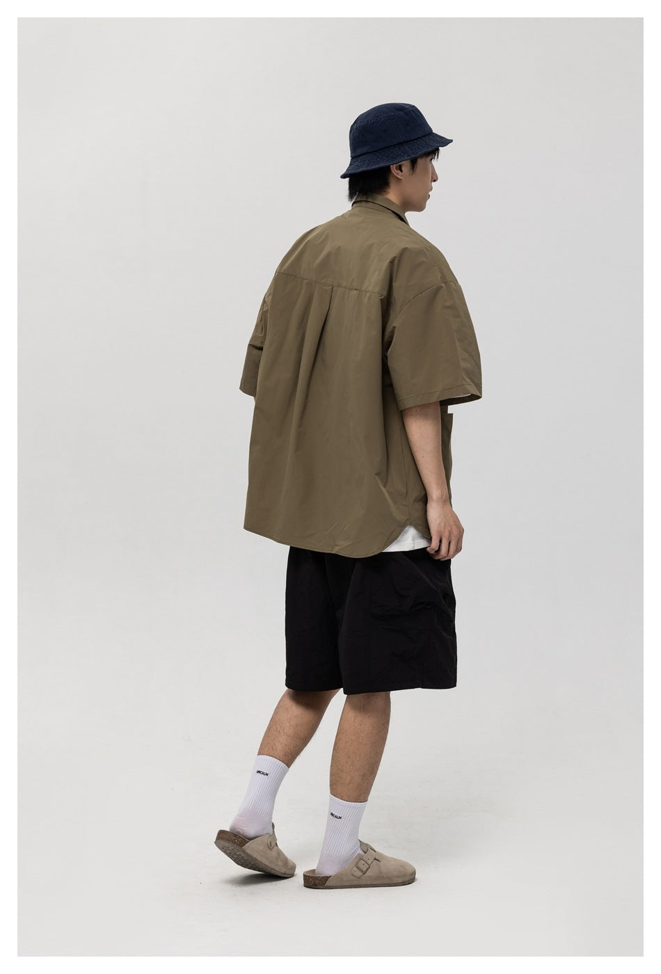 Model shown from behind wearing the olive short-sleeve shirt with black shorts and a navy bucket hat.