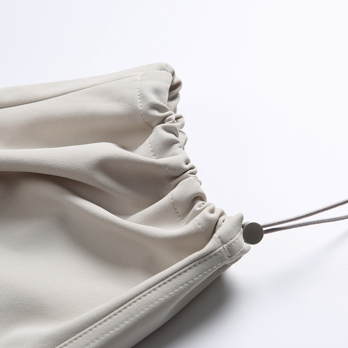 Close-up of a gathered trouser hem with a small drawcord toggle on light beige fabric.