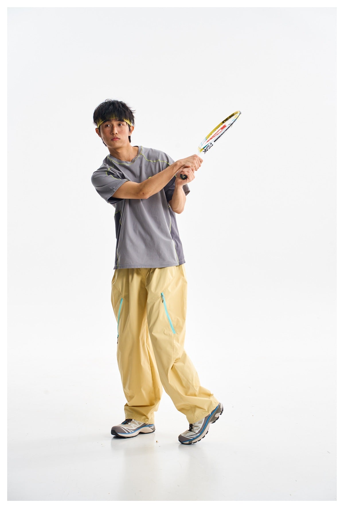 Model holding a tennis racket, wearing a gray color-block shirt with lime piping and loose pale yellow pants.