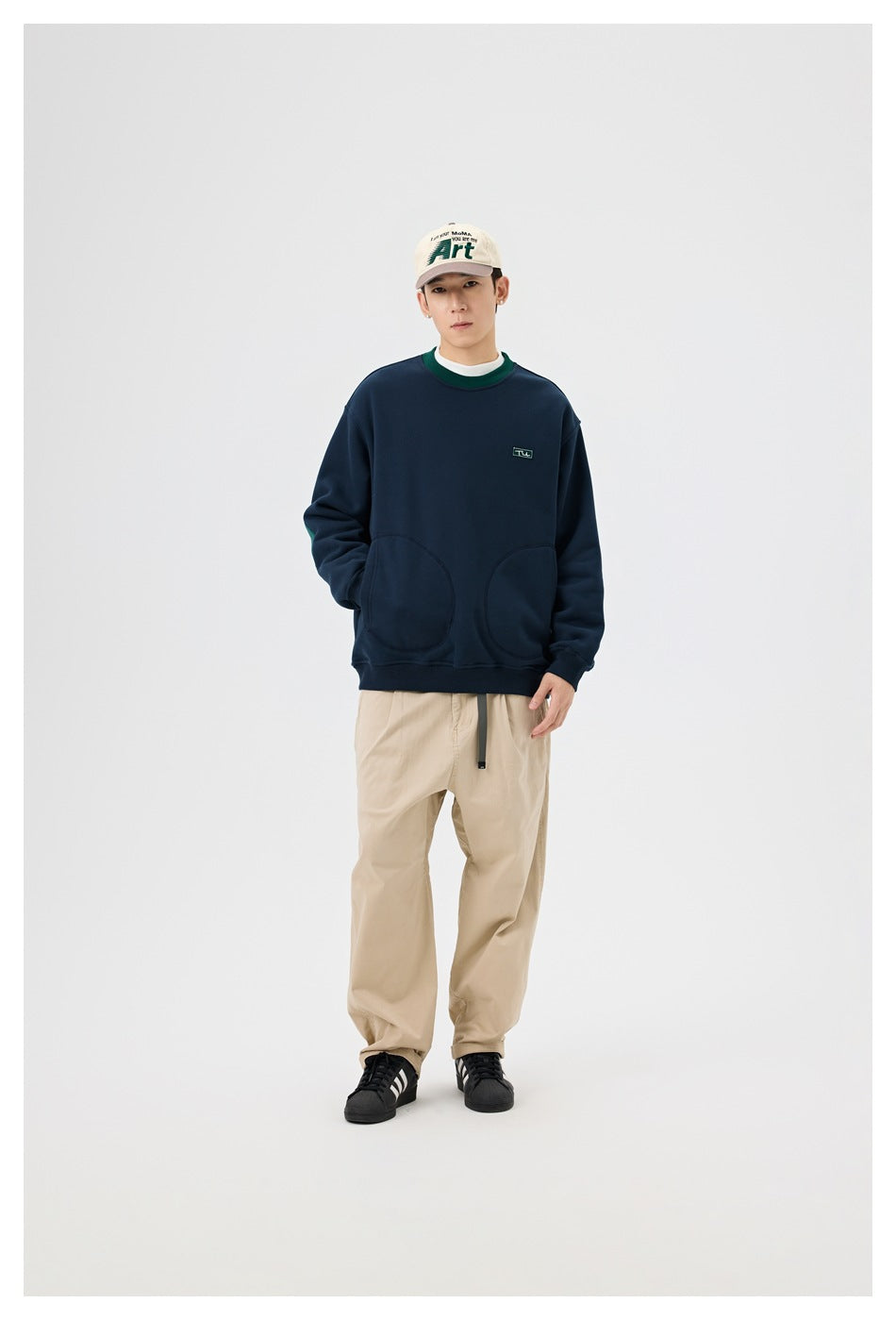 Full-body model wearing a navy sweatshirt with green collar trim and rounded front pockets, beige trousers, and a cream cap.