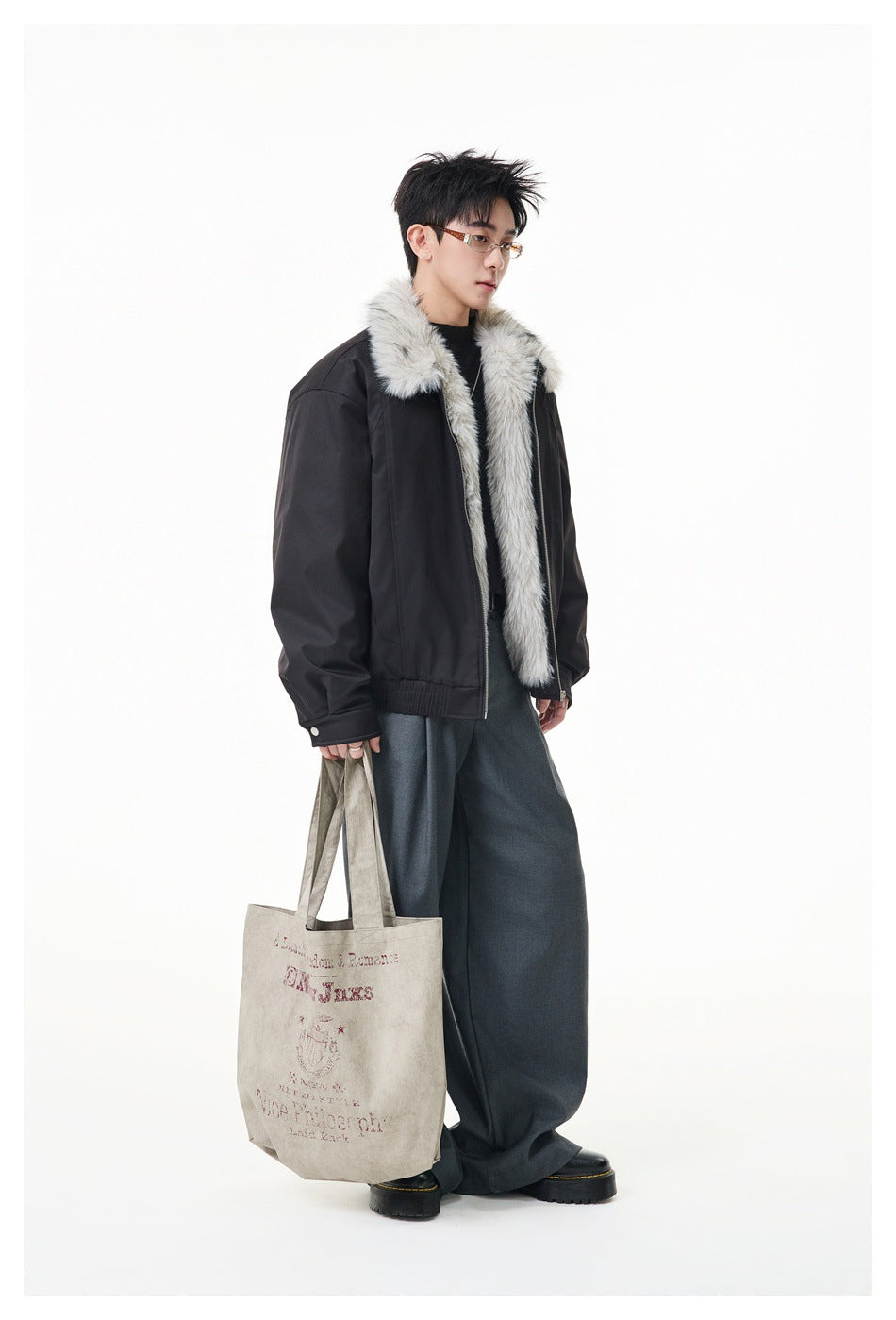 Model wearing open black jacket with fluffy gray and white collar and front trim, gray trousers and holding a tote bag