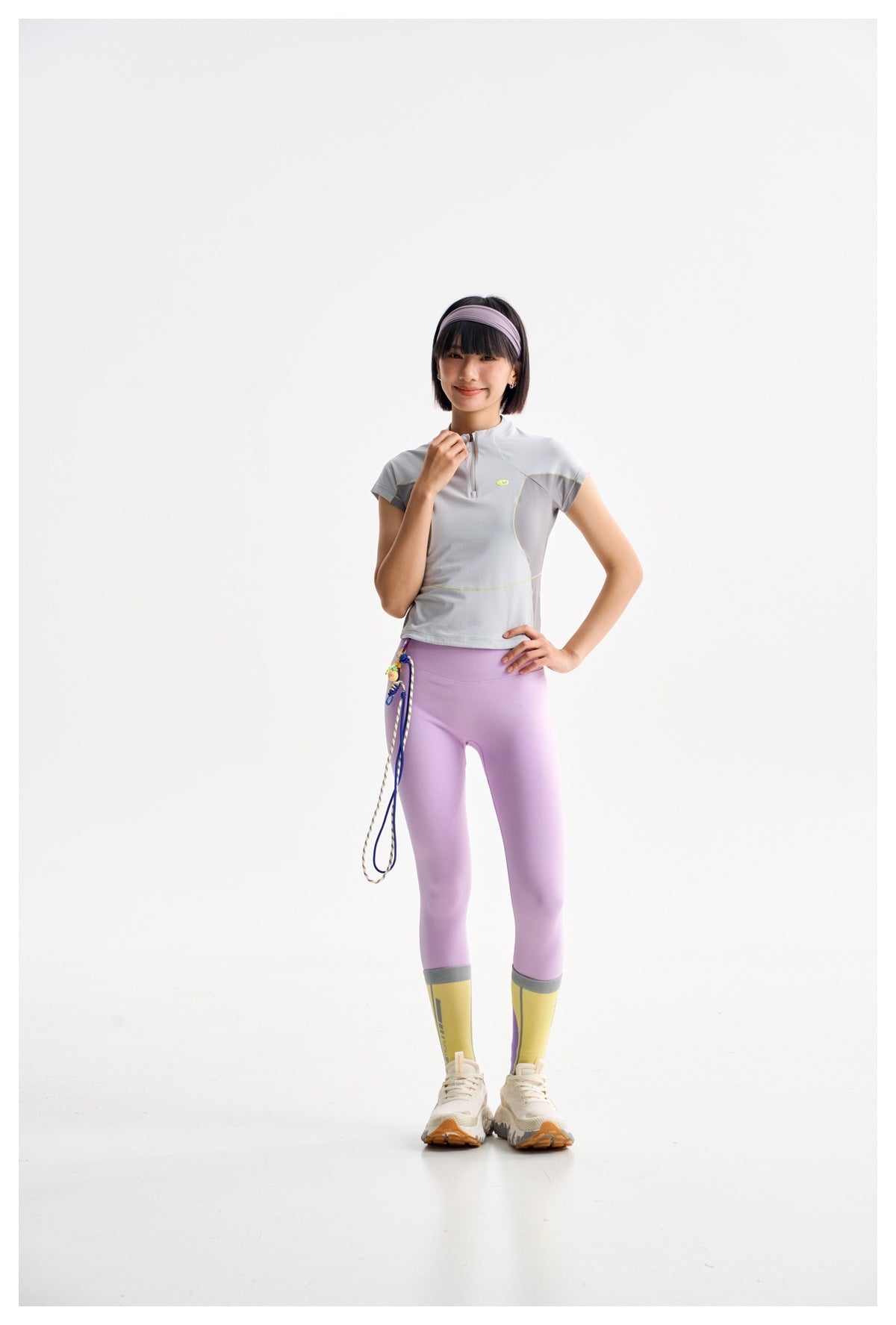 Model wearing a light gray athletic top with lavender leggings and yellow leg warmers, posing with one hand at the collar.