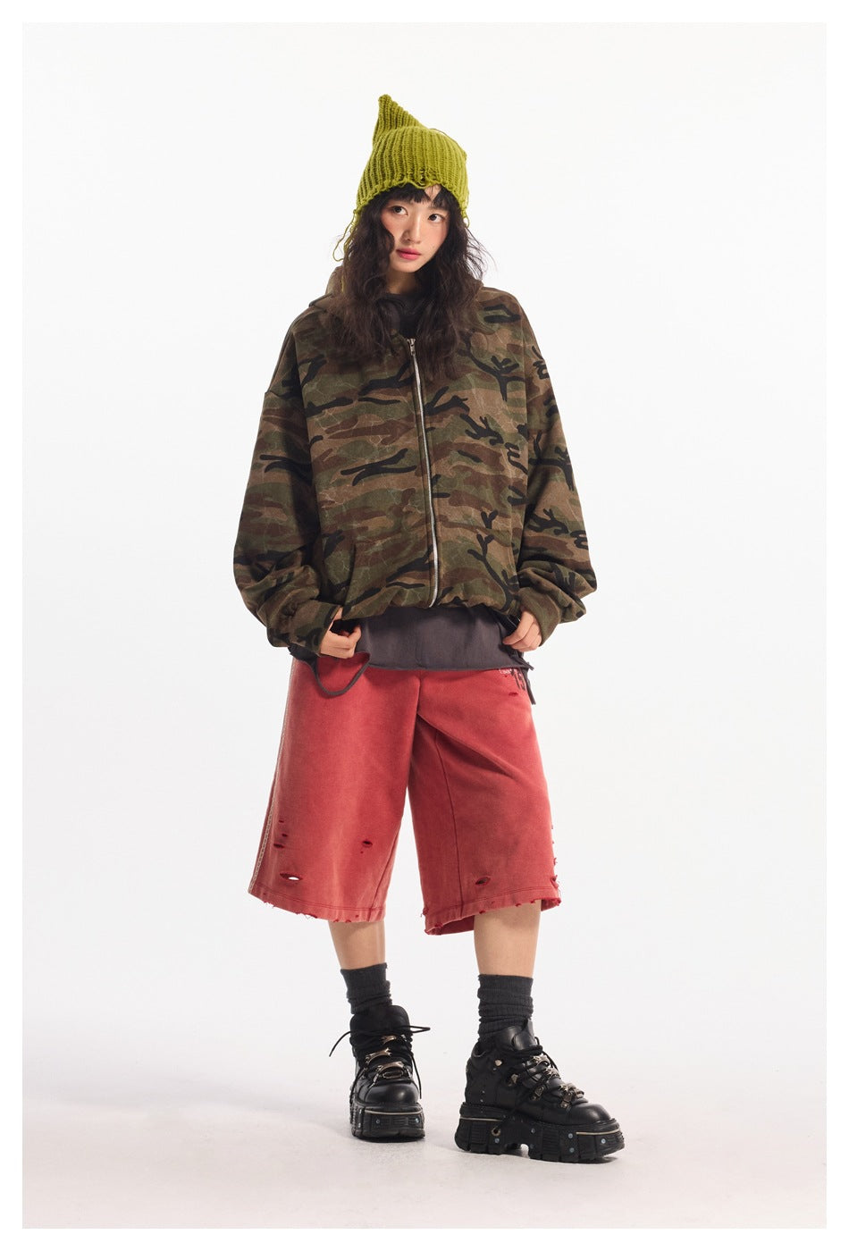 Model wearing an oversized camouflage zip hooded jacket with a green beanie, red shorts and black platform shoes