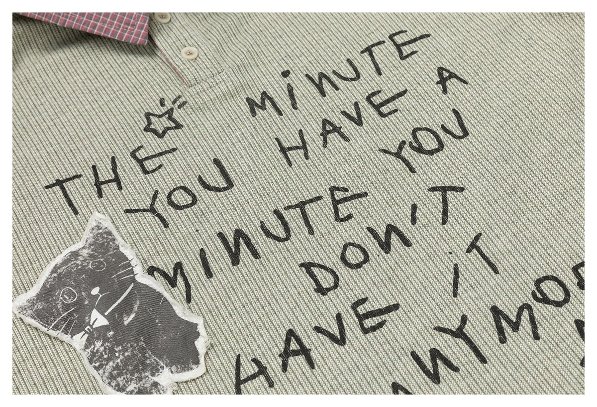 Sage-gray shirt close-up showing black handwritten-style lettering and a worn-looking cat patch.