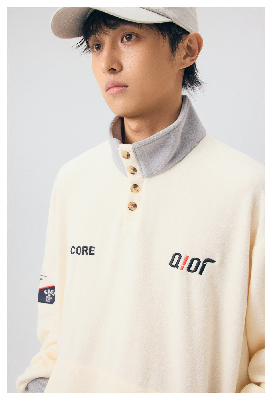 Model wearing a cream pullover with a gray stand collar, four-button placket, and embroidered chest details.