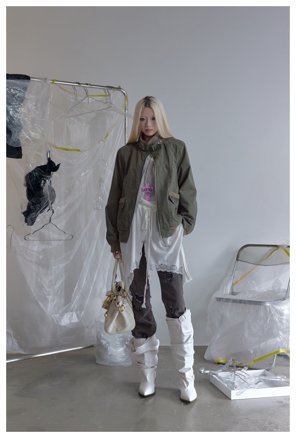 Model wearing the olive green stand-collar jacket open over a white layered top, with white boots and a cream handbag
