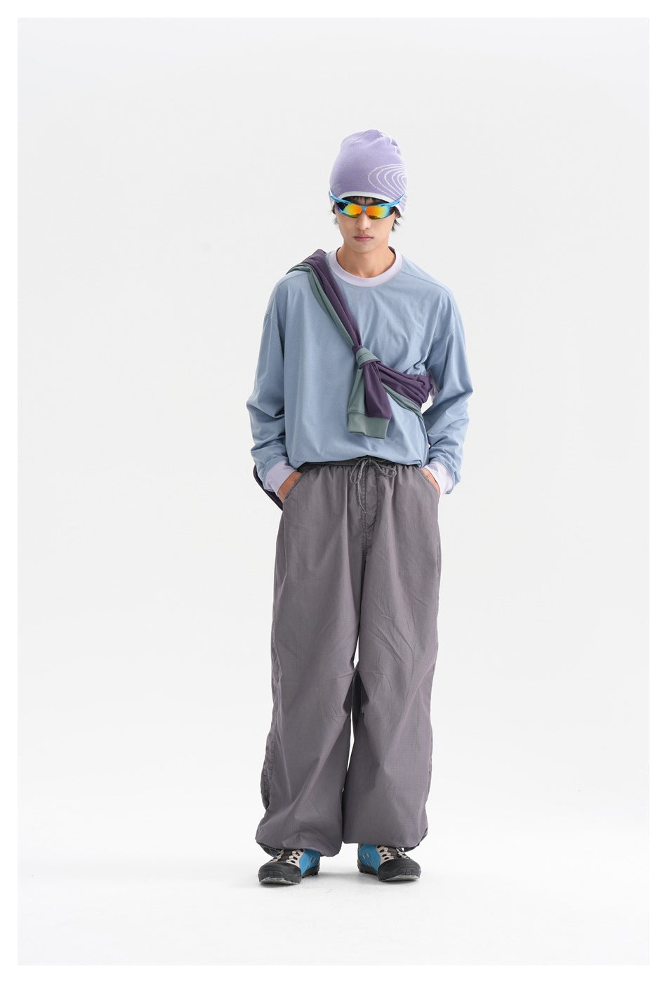 Person wearing a blue long-sleeve top with lilac collar and cuffs, gray wide pants, purple beanie and sunglasses