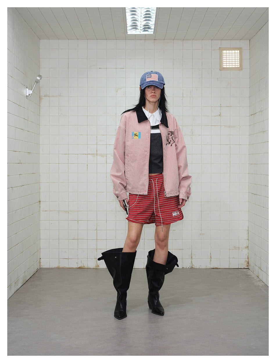 Model wearing the pink zip jacket open over a dark top and red striped shorts with a blue cap and black boots