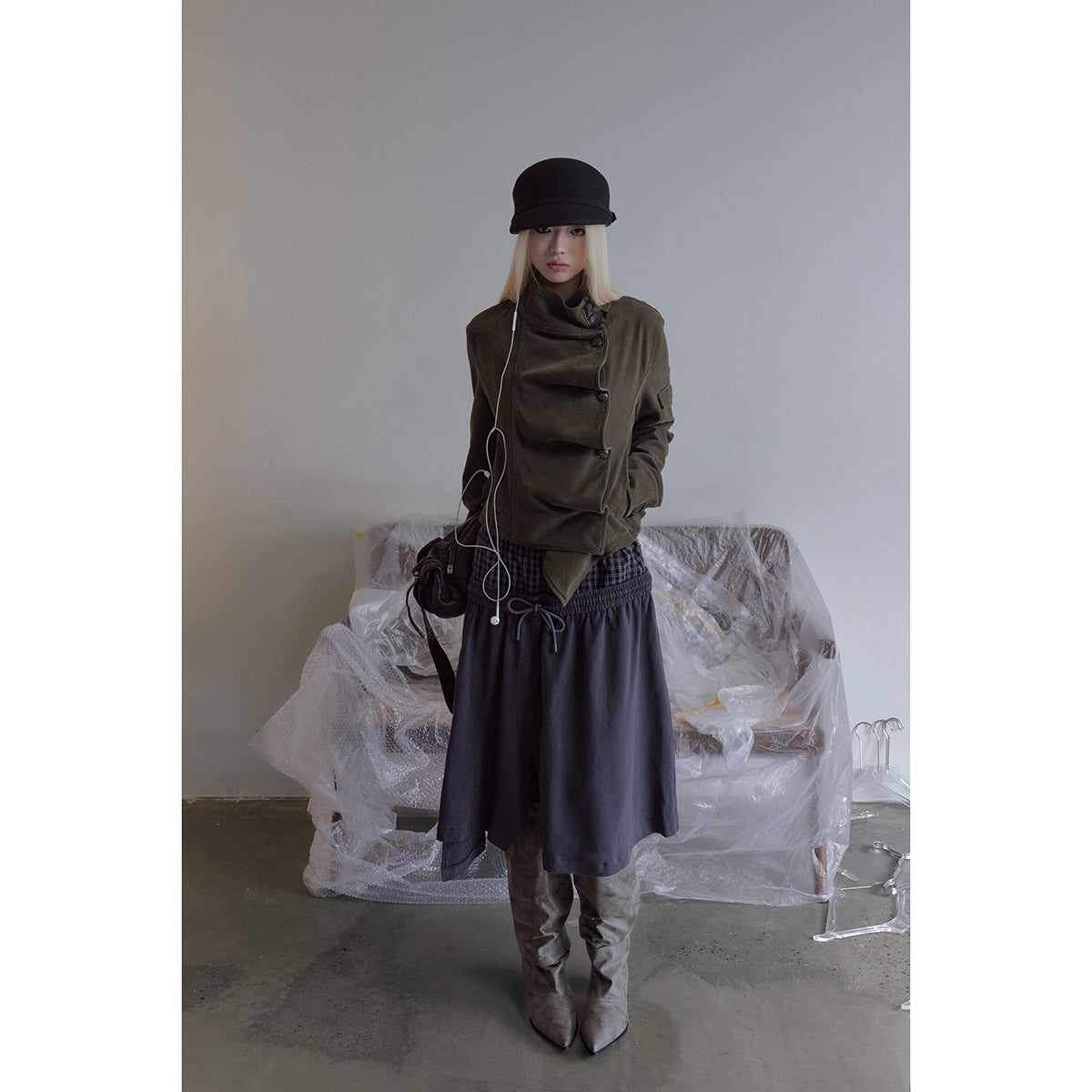 Full-length model in an olive asymmetrical jacket, dark skirt, black cap, and tall gray boots.