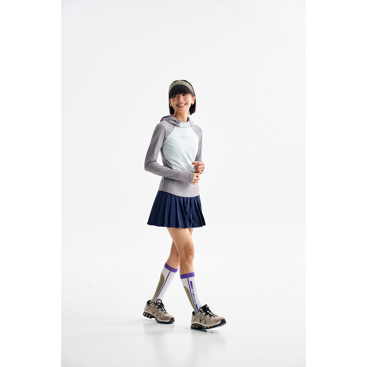 Model wearing a gray-and-white hooded top, navy pleated skirt, knee-high socks, and hiking sneakers.
