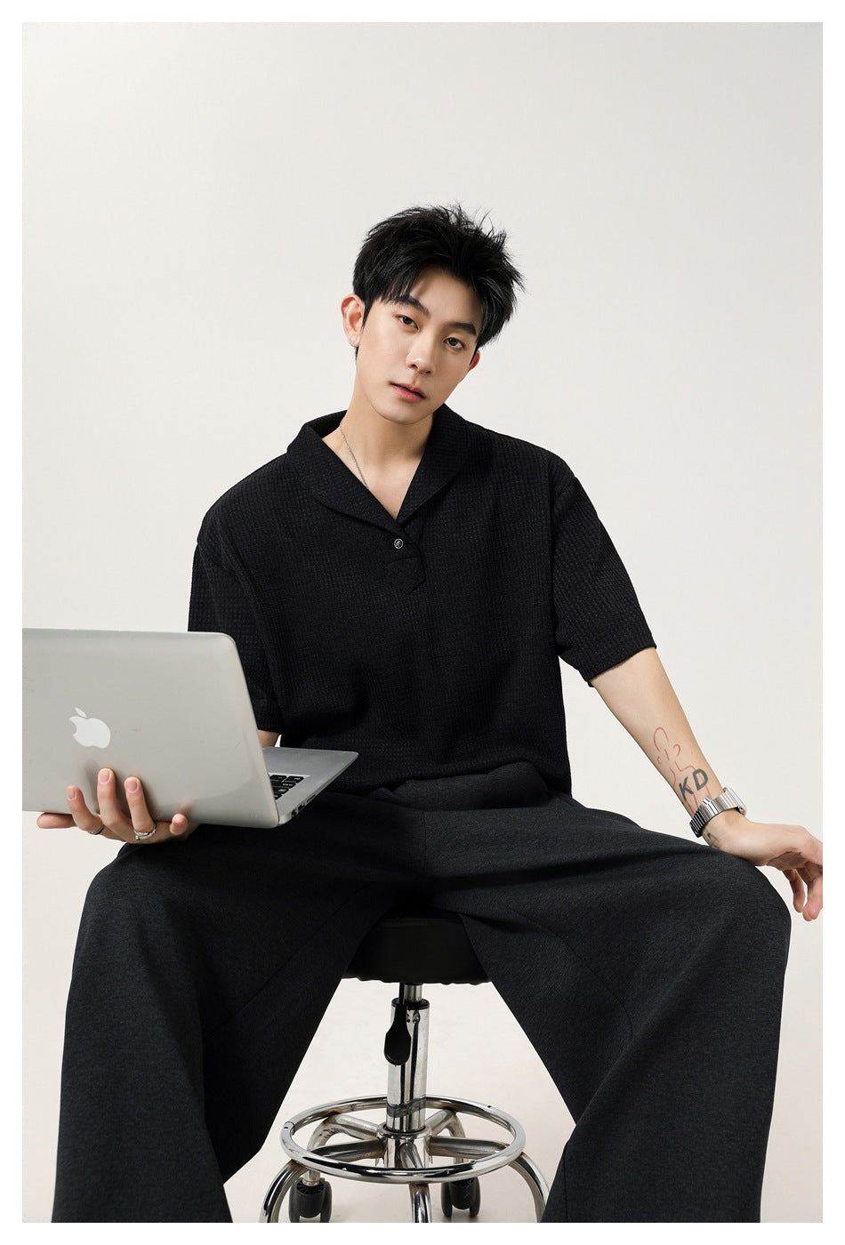 Seated model wearing a black textured polo with a notched collar and dark trousers.