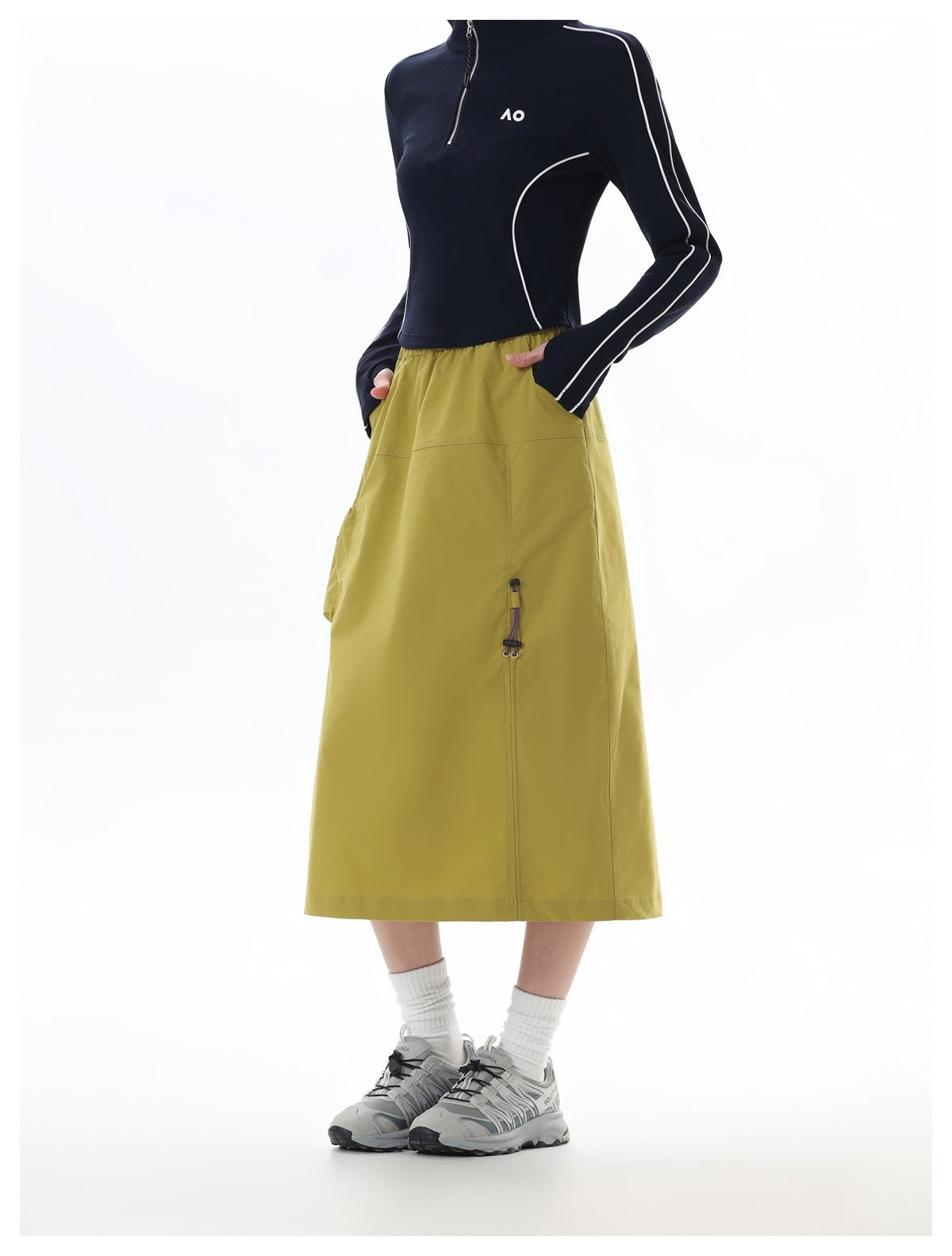 Model wearing a mustard-olive midi skirt with side pockets, paired with a navy zip-neck top with white piping.