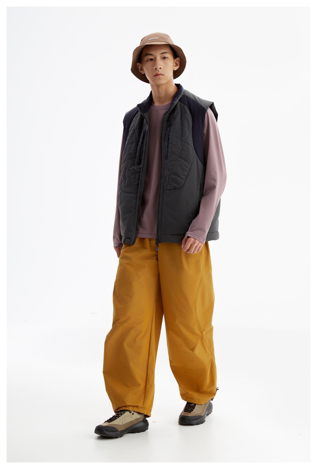 Model in a charcoal quilted vest with zip chest pockets, mauve long-sleeve shirt, mustard pants, and tan bucket hat.