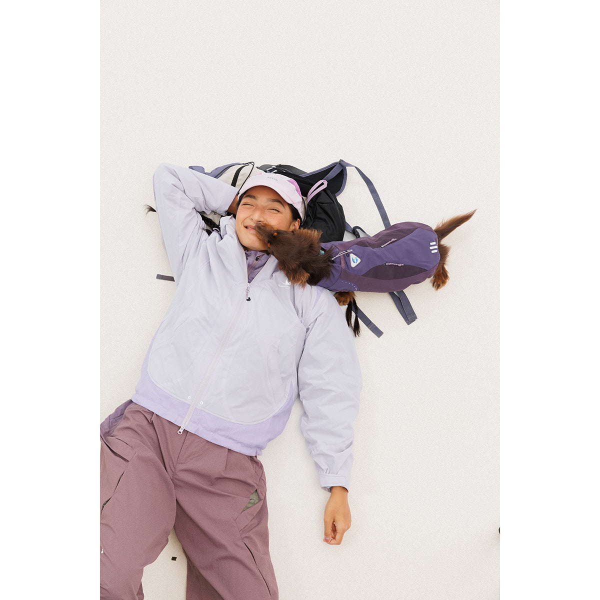Model reclining in a pale lavender hooded jacket and mauve hiking pants beside a purple backpack and small dog.