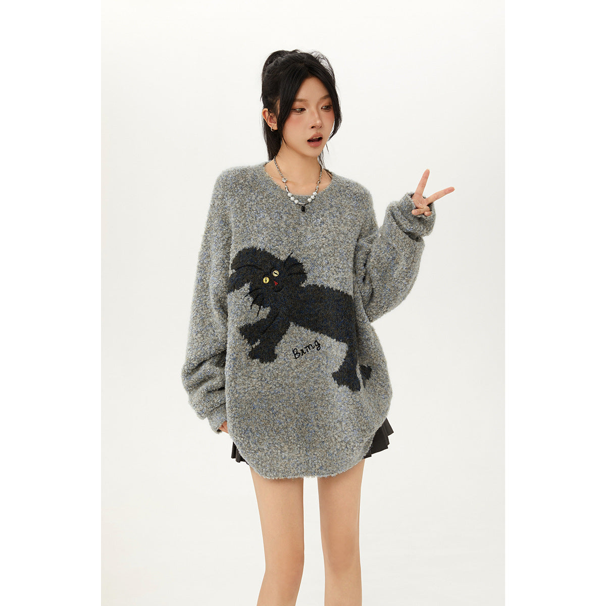 Model wearing a gray-blue cat-graphic sweater, raising a peace sign; a black skirt peeks out below.