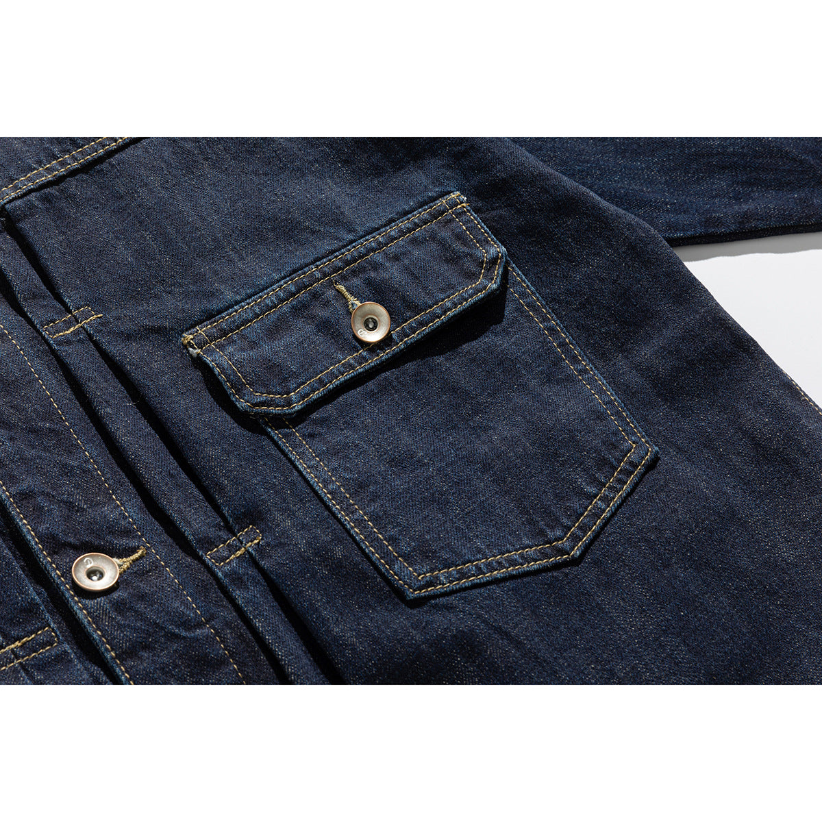 Close-up of a dark indigo denim jacket's buttoned flap pocket and golden contrast stitching.