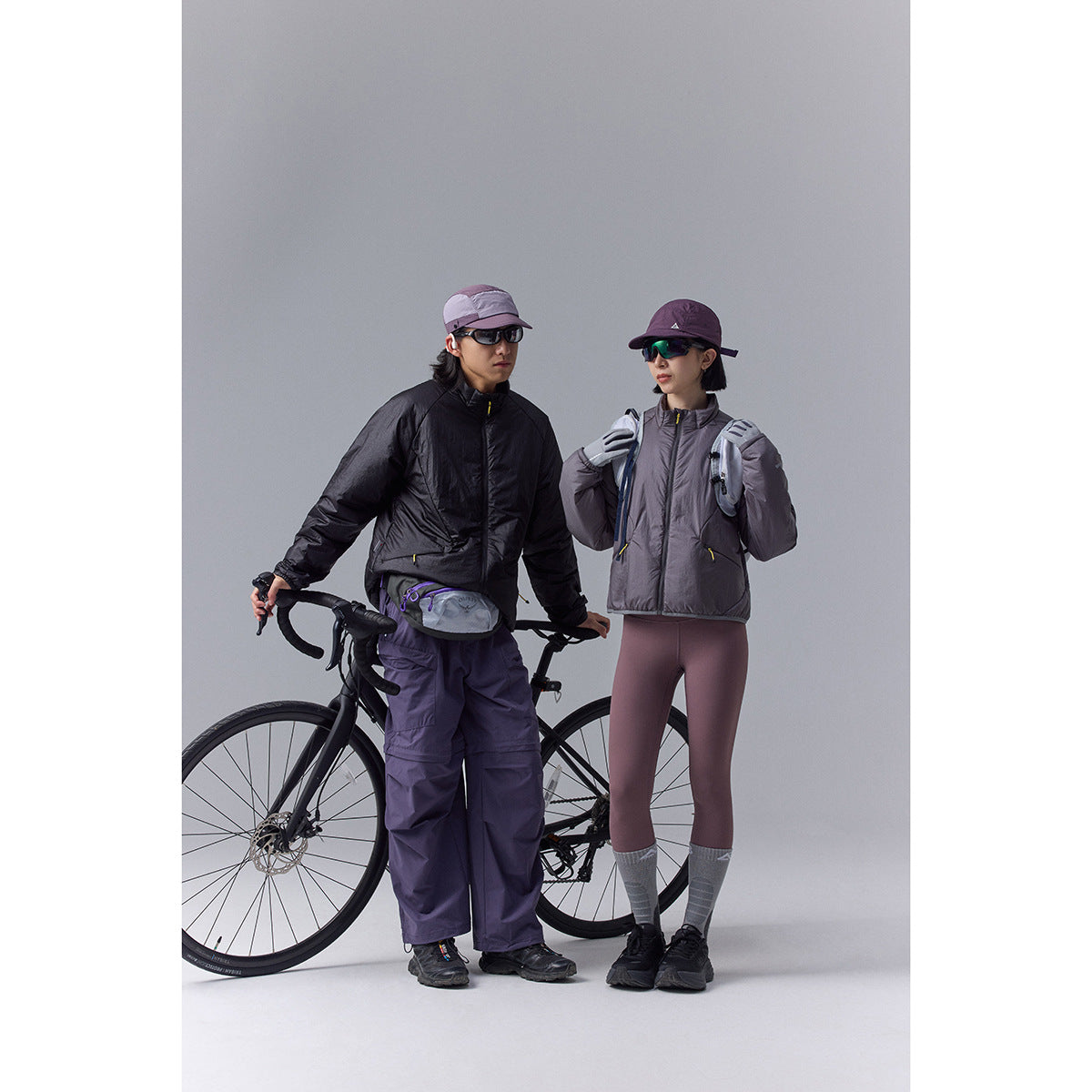 Two cyclists wear black and gray-purple jackets with outdoor pants and caps beside a road bike.