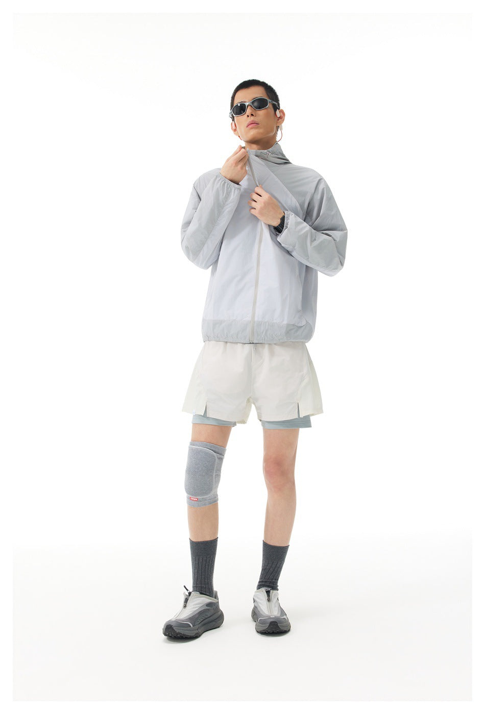 Model wearing a light gray hooded zip jacket with white shorts, gray knee support, dark socks, and sneakers.