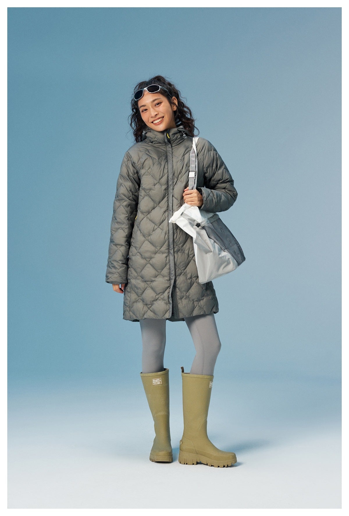 Full-length view of a model wearing a gray quilted hooded coat with gray leggings and olive boots.
