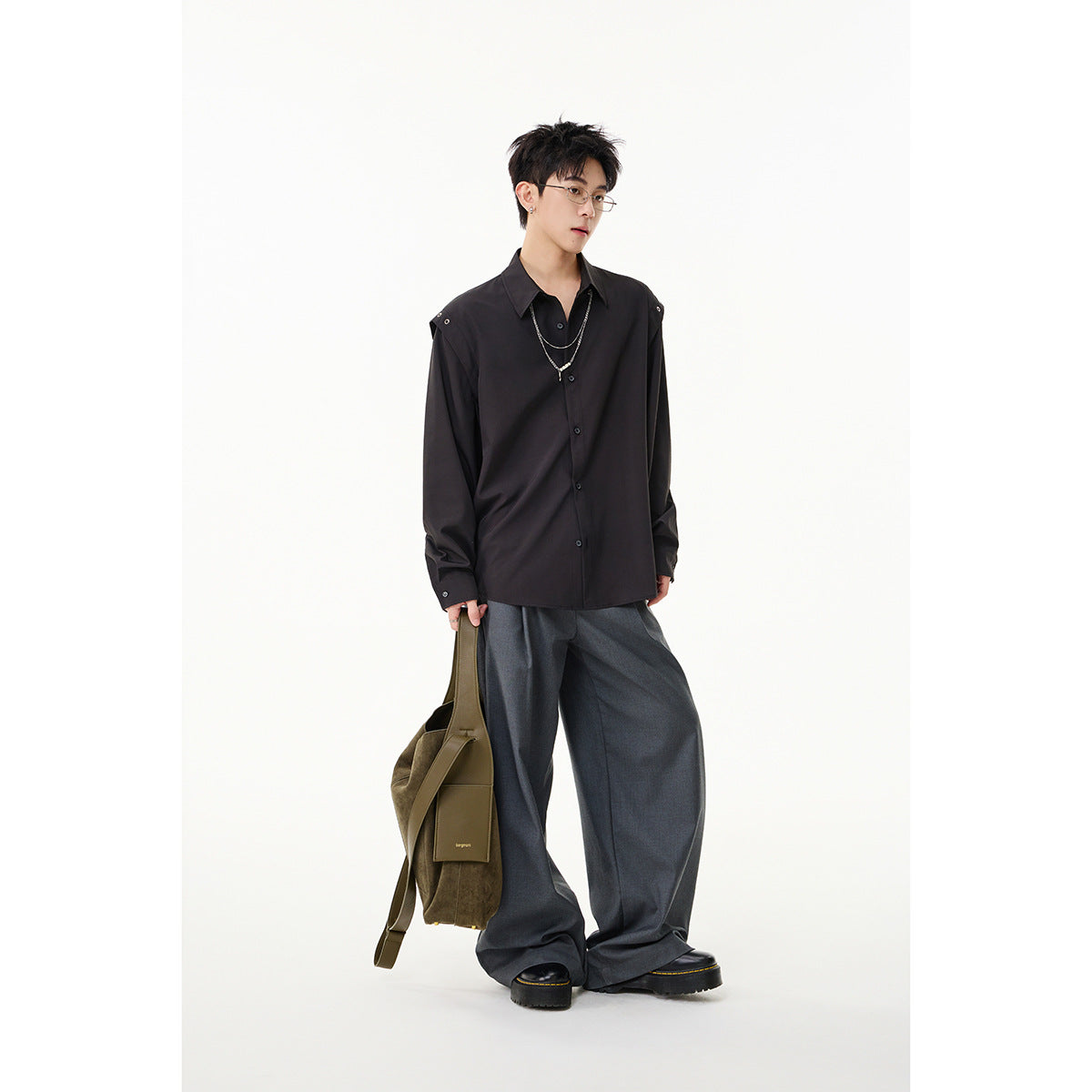 Model wearing a black button-front shirt with shoulder snaps and layered chains, paired with wide gray trousers.