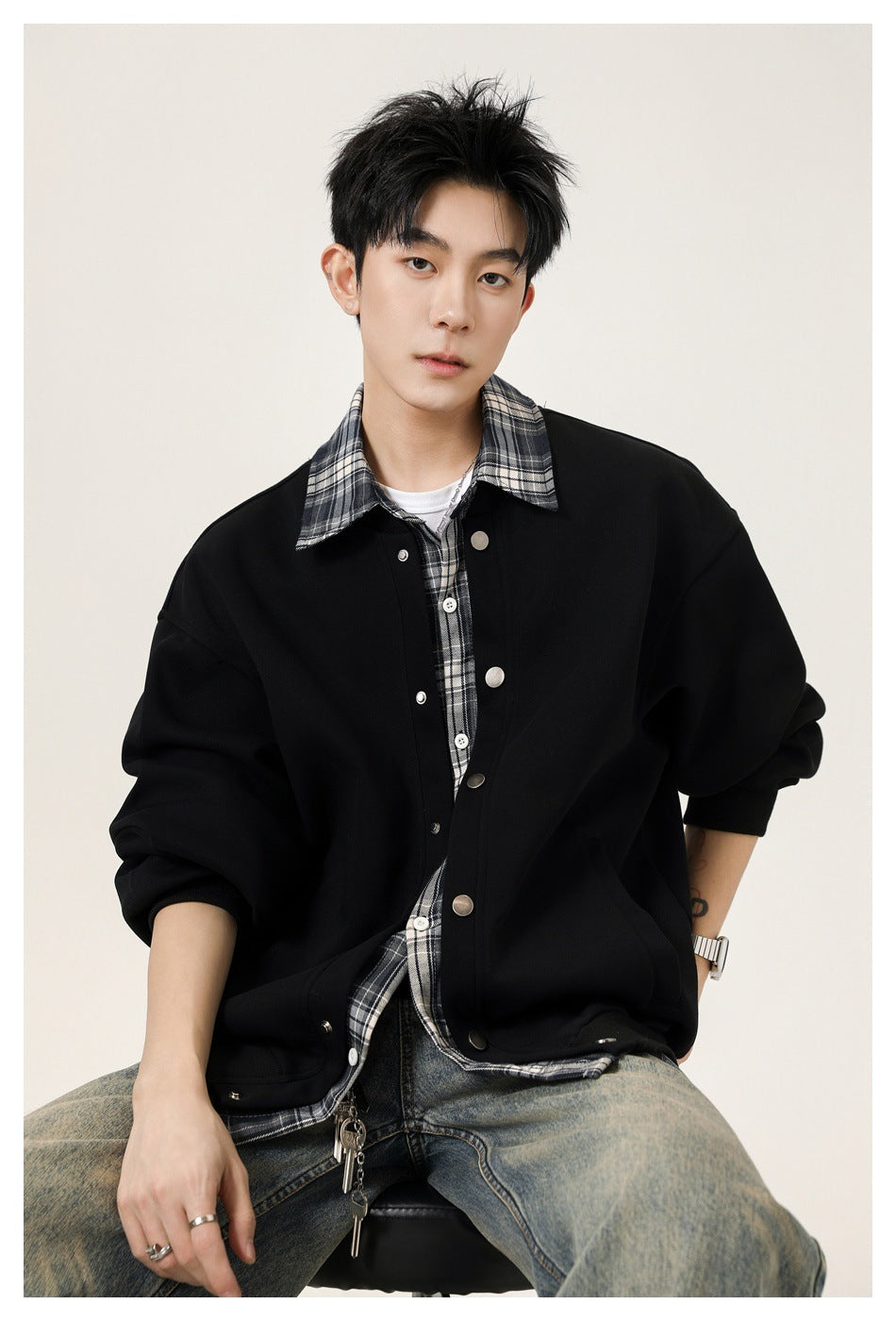 Seated model wearing an oversized black snap-front jacket with a gray plaid collar and shirt.