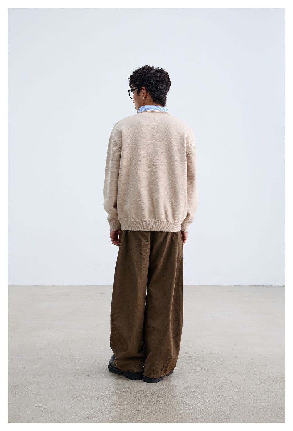 Rear view of a model wearing a beige sweater over a striped shirt with loose brown trousers.