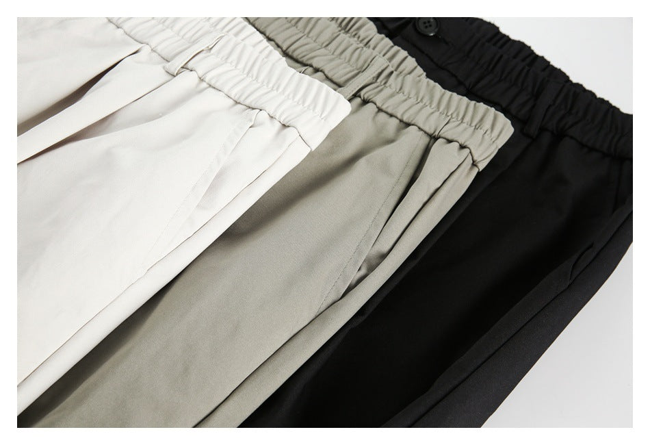 Close view of off-white, gray-khaki, and black trousers overlapping, showing gathered waistbands and front pleats.