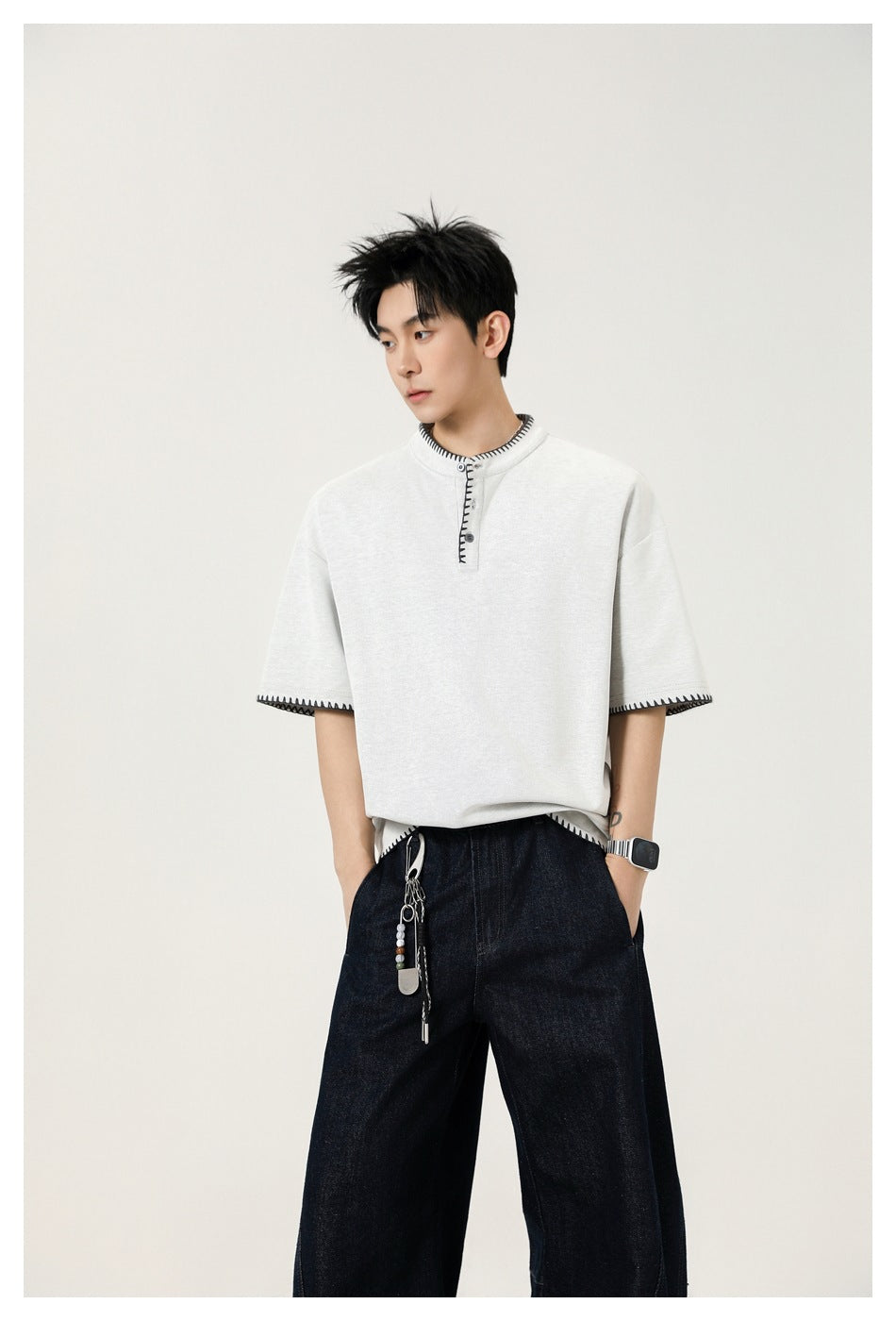 Man wearing light gray Henley top with dark stitched trim, dark wide-leg jeans and a keychain, hands in pockets