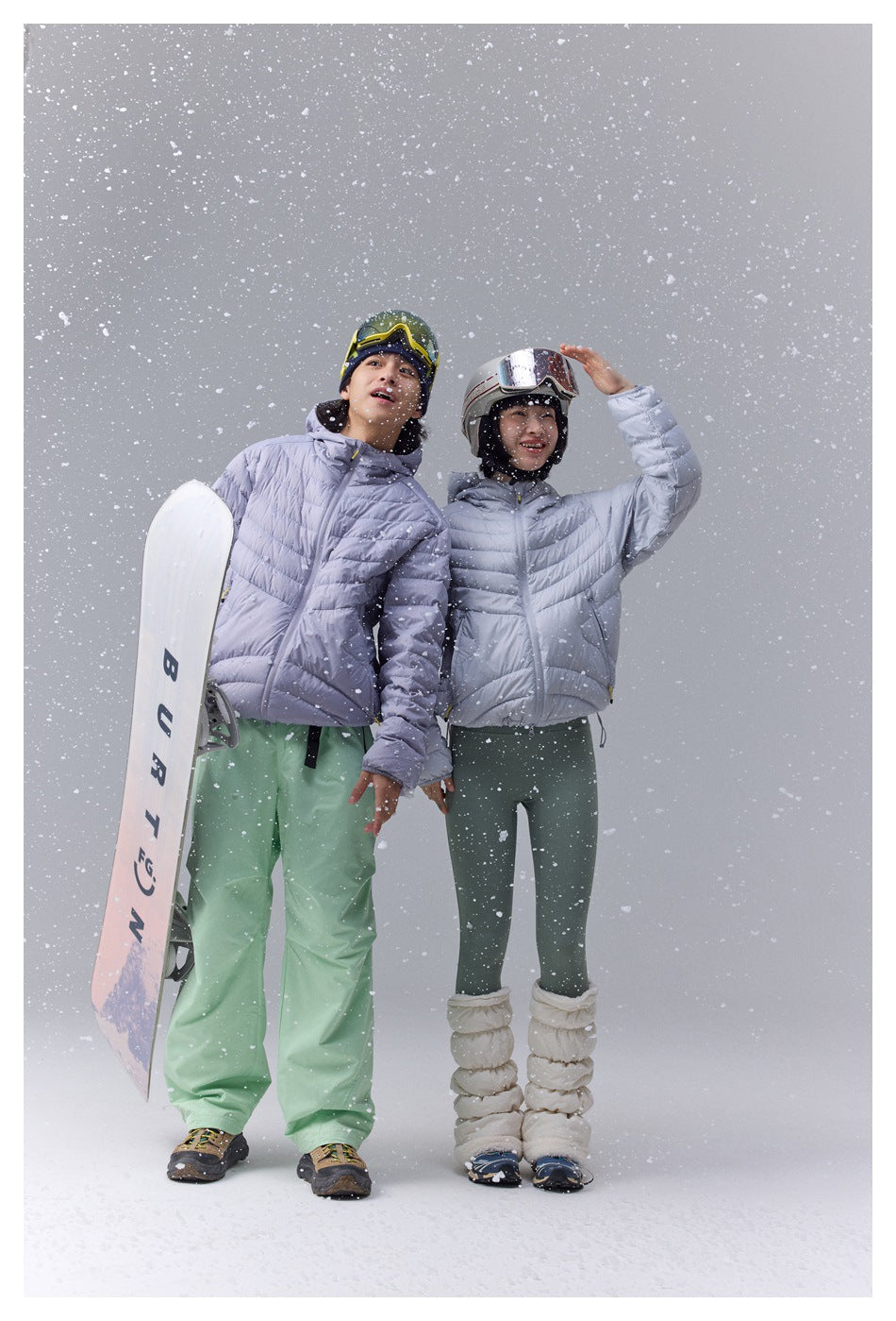 Two models in pale lavender puffer jackets pose with a snowboard amid falling snow.