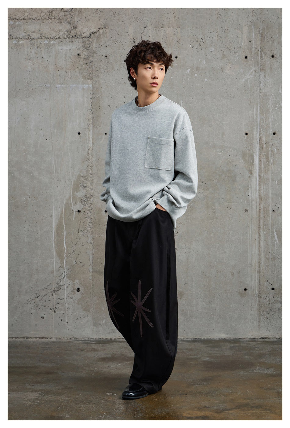 Model wearing a light gray oversized crewneck sweatshirt with a chest pocket and black trousers with starburst details.