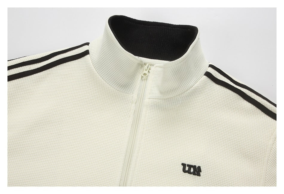 Close-up of an ivory zip-up jacket collar, zipper, black shoulder stripes, and embroidered chest logo.