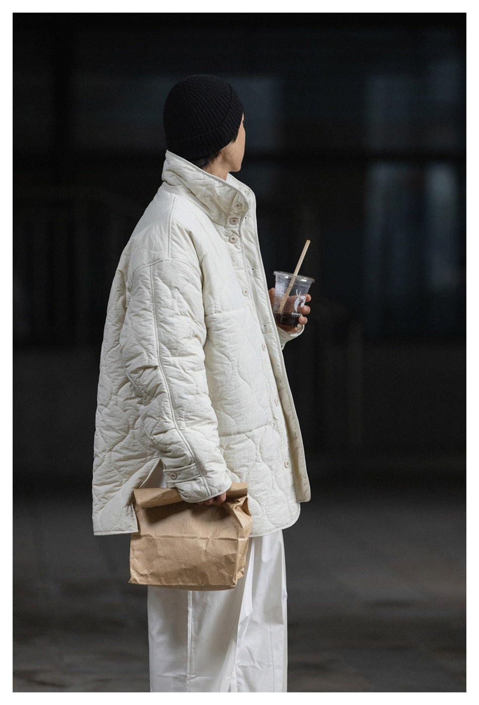 Person wearing an off-white quilted jacket with white trousers, black beanie, holding a drink and paper bag