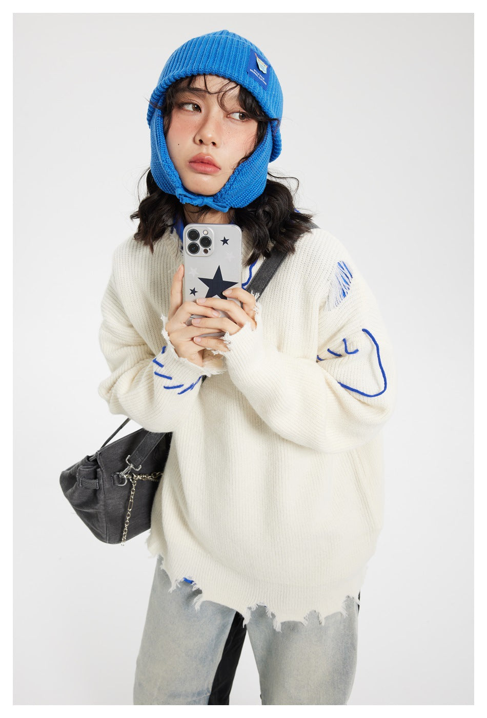Model wearing an oversized ivory distressed sweater with blue stitched accents and a bright blue knit earflap hat.