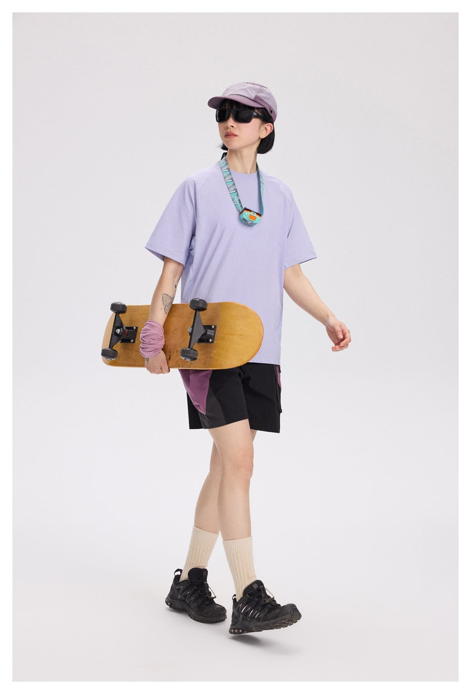 A woman models a loose lavender short-sleeve T-shirt with black shorts, a mauve cap, and sunglasses.
