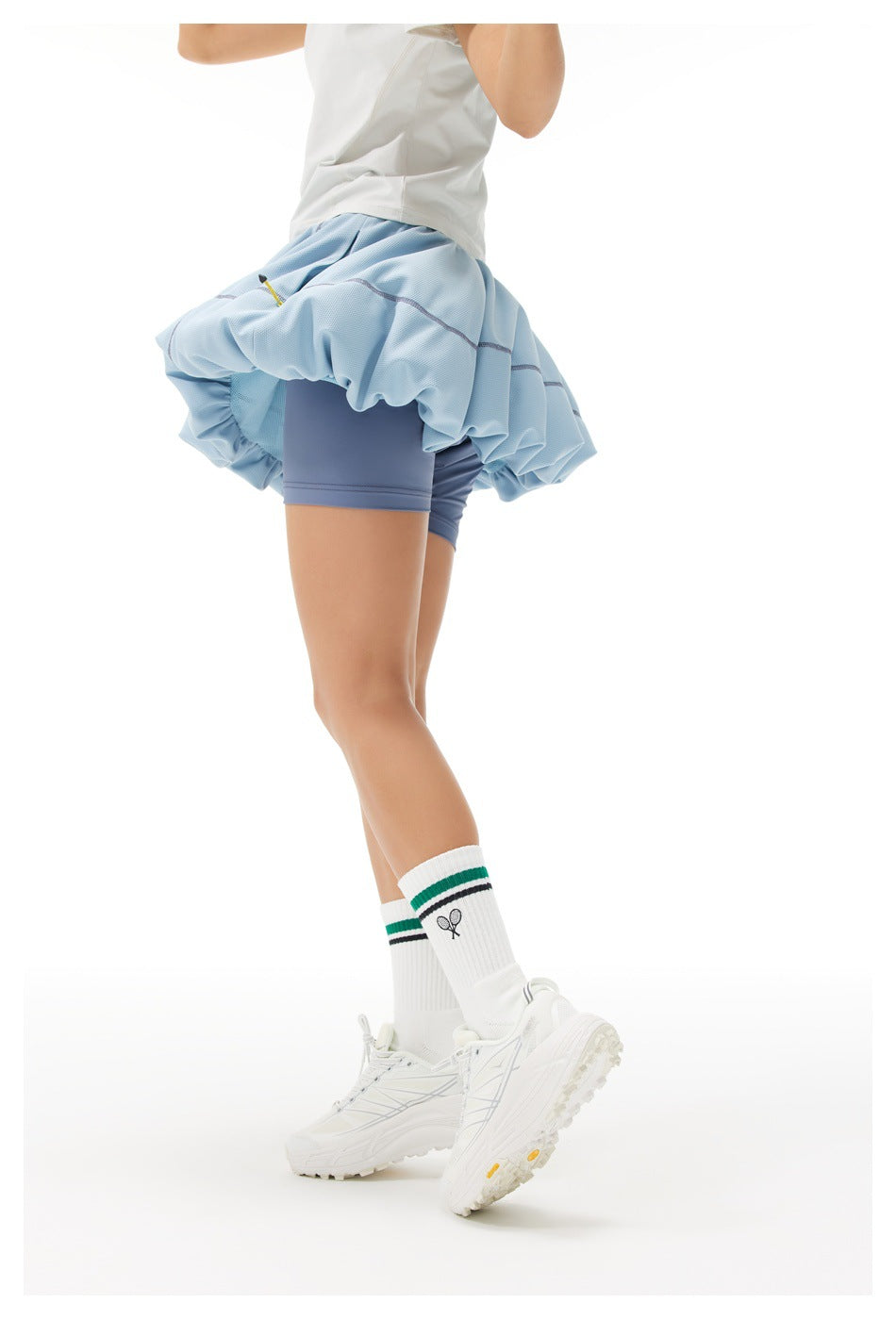 Rear side view of a model wearing a pale blue bubble tennis skirt over slate-blue shorts.