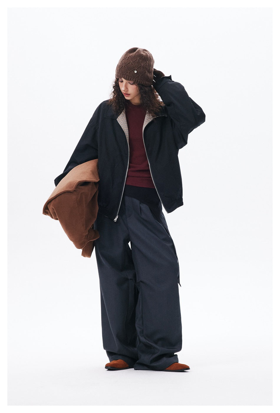 Model in a black checked-lining jacket and loose dark trousers, touching her brown knit beanie; full outfit visible.