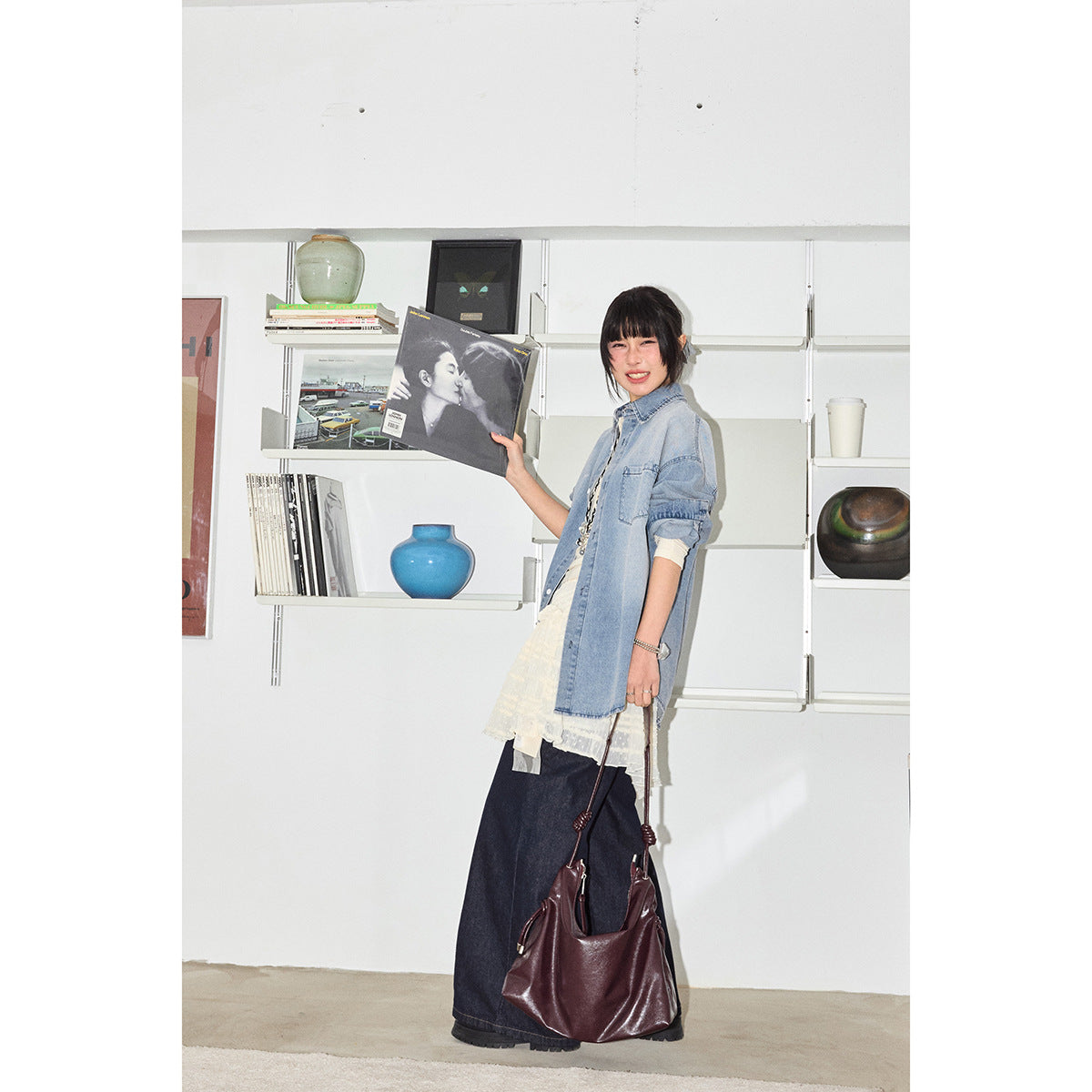 Standing model in an oversized light blue denim shirt over cream layers and dark wide-leg jeans, holding a burgundy bag.