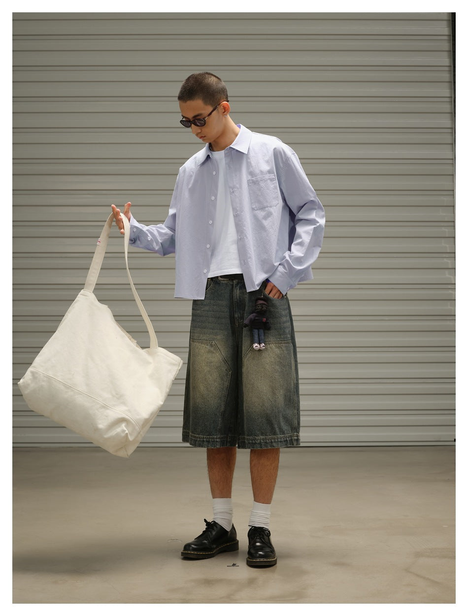 Full-length view of a model in a pale blue button-up shirt and oversized dark denim shorts, carrying a cream tote.