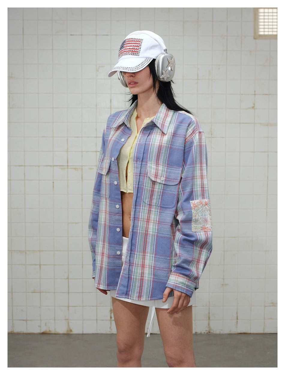 Model wearing a blue plaid button-up shirt with a floral sleeve patch, pale yellow cropped top, white skirt, cap and