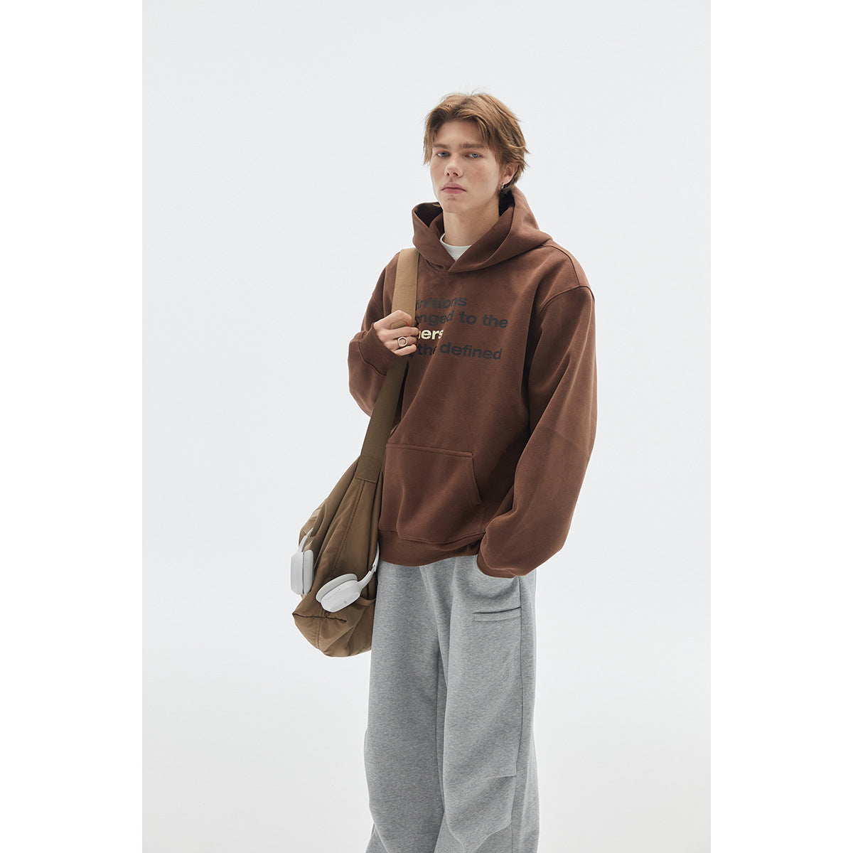 Model wearing a brown slogan-print pullover hoodie with a kangaroo pocket and gray sweatpants.