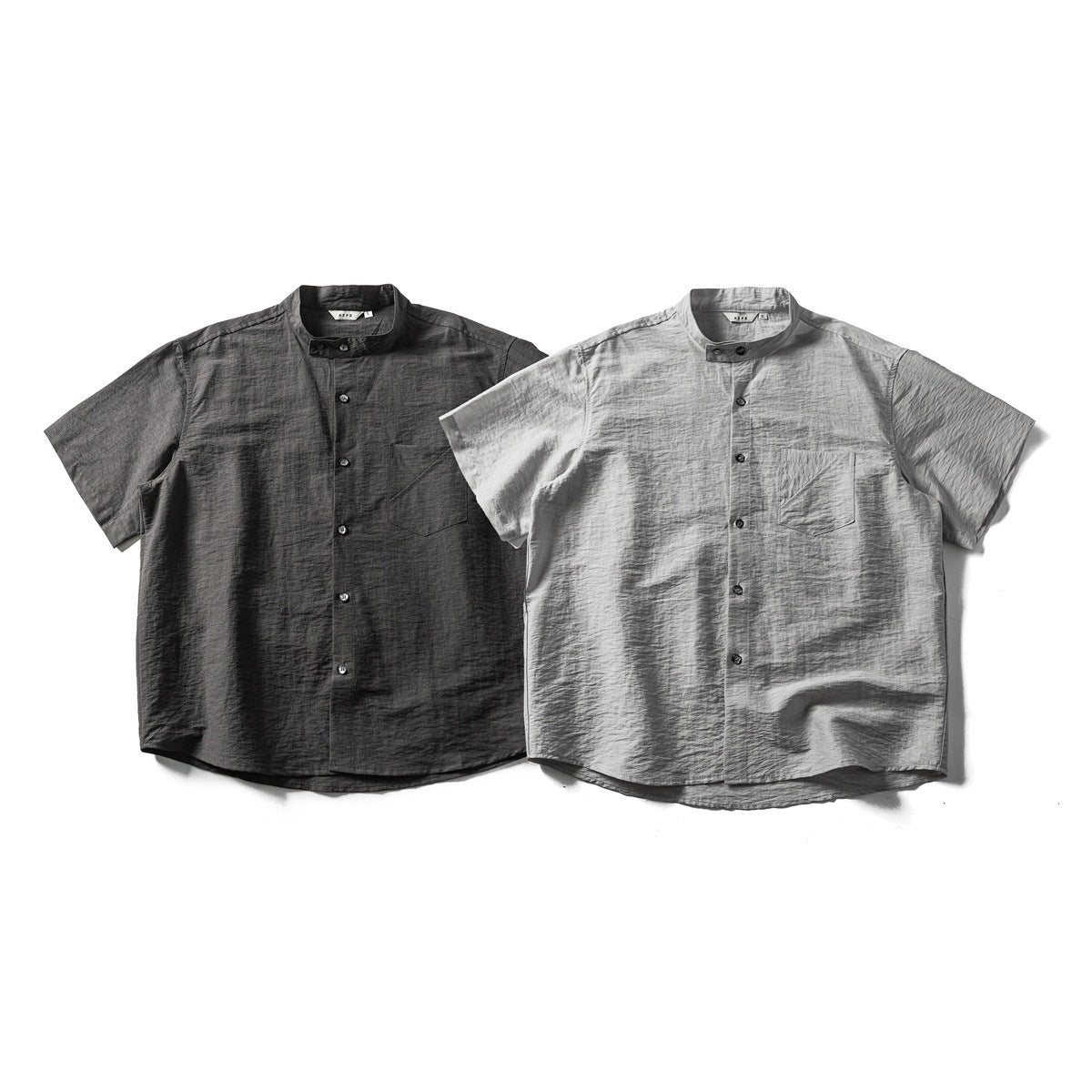 Two short-sleeved band-collar button-up shirts shown from the front, one charcoal and one light gray, each with a chest